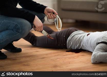 murder and robbery concept - criminal or murderer holding knife in blood and jewelry and dead woman body lying on floor at crime scene (staged photo). criminal with knife and jewelry at crime scene