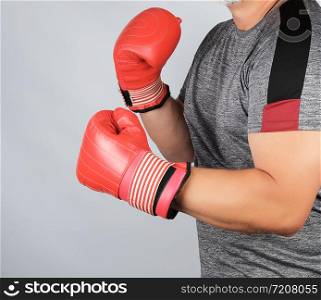 muscular adult athlete in gray uniform and red leather boxing gloves standing in a rack, white background