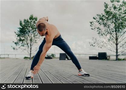 Muscular athlete does sport exercises, warms up body before cardio training, poses outdoor, dressed in active wear, leads active lifestyle. Sport and fitness concept. Bodybuilder with perfect biceps