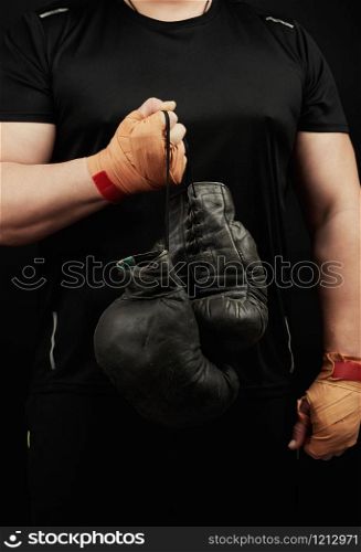 muscular athlete in a black uniform holds very old black boxing gloves in his hand, his hands are bandaged with an orange elastic sports bandage, black background