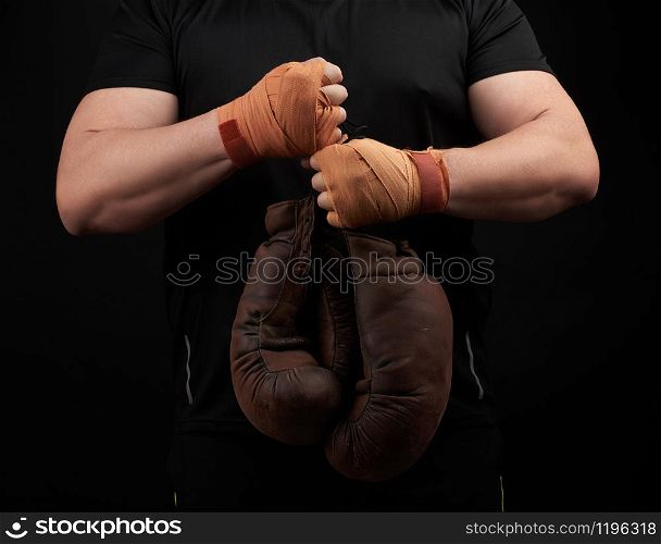 muscular athlete in a black uniform holds very old brown boxing gloves in his hand, his hands are bandaged with an orange elastic sports bandage, black background