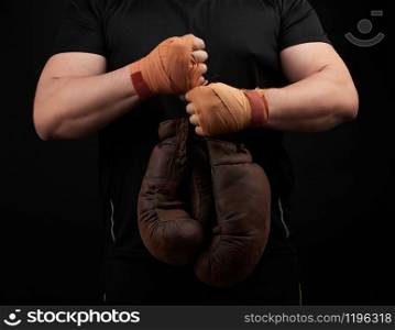 muscular athlete in a black uniform holds very old brown boxing gloves in his hand, his hands are bandaged with an orange elastic sports bandage, black background