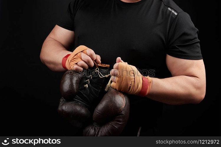 muscular athlete in a black uniform holds very old brown boxing gloves in his hand, his hands are bandaged with an orange elastic sports bandage, black background, copy space
