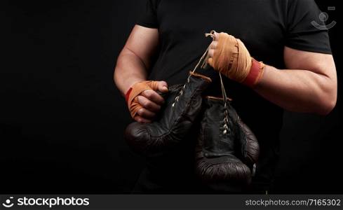 muscular athlete in a black uniform holds very old brown boxing gloves in his hand, his hands are bandaged with an orange elastic sports bandage, black background, copy space