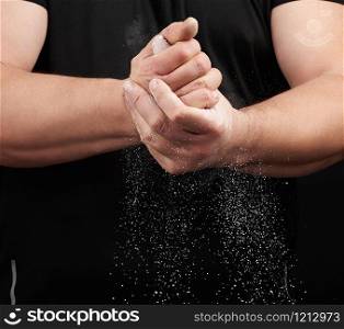 muscular athlete in a black uniform rubs his hands with white dry sports magnesia, powder scatters in different directions on a black background