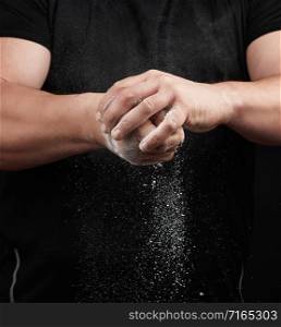 muscular athlete in a black uniform rubs his hands with white dry sports magnesia, powder scatters in different directions on a black background