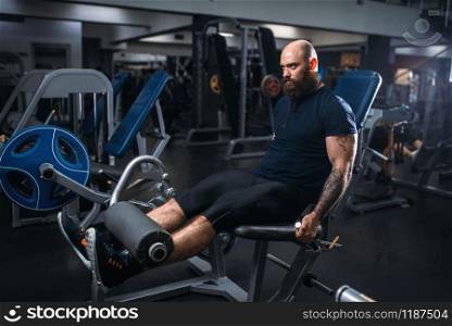 Muscular athlete trains legs on exercise machine, training in gym. Bearded man on workout in sport club, healthy lifestyle. Muscular athlete trains legs on exercise machine