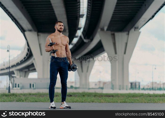 Muscular athletic bearded bodybuilder raises dumbbells works on biceps has hard workout near bridge dressed in sport trousers and sneakers, being motivated, enjoys sport. Healthy lifestyle concept