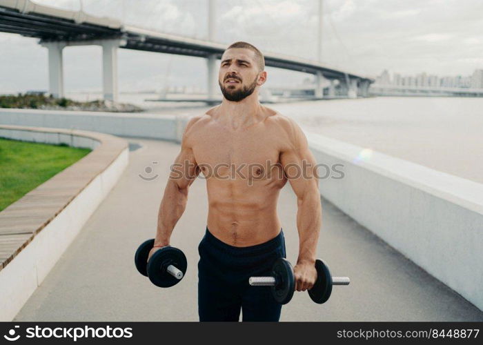 Muscular bodybuilder concentrated into distance has strong body strong muscles holds barbells and does exercises for biceps outdoor poses near river with bridge. Healthy lifestyle and sport concept