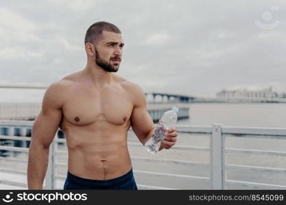 Muscular build bearded man goes in for sport, holds bottle of fresh water, focused into distance, poses on bridge near river during cloudy day, takes break after physical exercises. Cardio workout