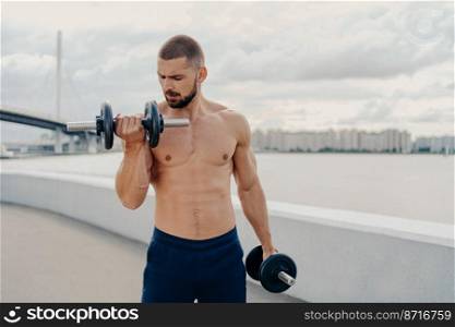 Muscular guy with naked torso exercises with barbells, stands shirtless, trains muscles outdoor, stands on piere, has motivation to keeping fit and healthy. Fitness, sport, bodybuilding concept