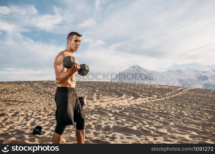 Muscular male athlete doing exercises with dumbbells in desert at sunny day. Strong motivation in sport, strength outdoor training, sportsman with weights