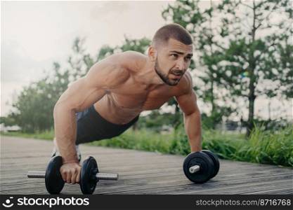 Muscular male bodybuilder does push up with barbells stands in palnk pose with naked body, trains determination, goes in for sport regularly. Sport, fintess, powerlifting and bodybuilding concept
