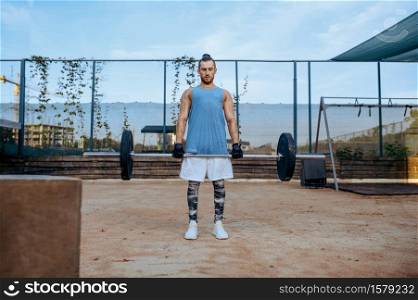 Muscular man does an exercise with a barbell, street workout. Fitness training on sports ground outdoor, male person pumps muscles, active urban lifestyle. Man does an exercise with barbell, street workout