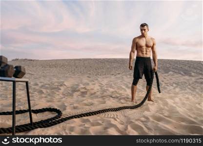Muscular man doing exercise with rope in desert at sunny day. Strong motivation in sport, strength outdoor training. Muscular man doing exercise with rope in desert
