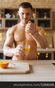 Muscular man with naked body cooking orange juice on the kitchen. Nude male person preparing breakfast at home, food preparation without clothes