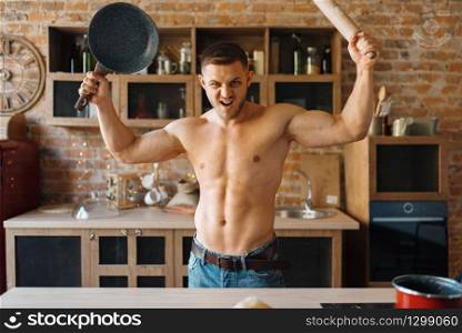 Muscular man with naked body holds frying pan and rolling pin on the kitchen. Nude male person preparing breakfast at home, food preparation without clothes