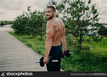 Muscular satisfied athlete bearded man gets energy during sport training, has strong arms, raises barbells, dressed in shorts, poses outdoor, enjoys regular workout in nature, builds healthy body.