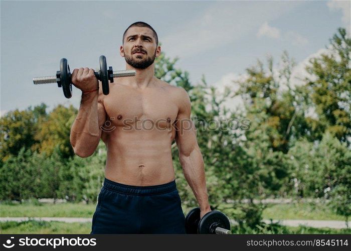 Muscular shirtless young man raises barbells outdoors, trains muscles and has strong body. Athletic sportsman with strong arms leads healthy lifestyle, enjoys training against nature background.