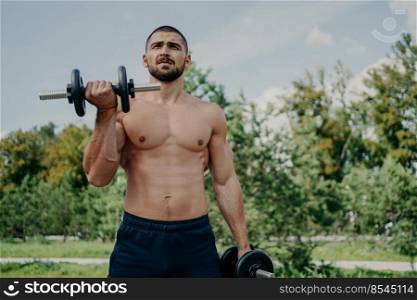 Muscular shirtless young man raises barbells outdoors, trains muscles and has strong body. Athletic sportsman with strong arms leads healthy lifestyle, enjoys training against nature background.