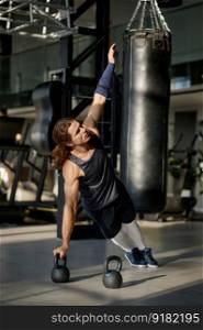Muscular sportsman doing cross training kettlebell routine at fitness club. Athletic man standing in plank with raised up hand and pressing on kettle bell. Muscular sportsman doing cross training kettlebell routine at fitness club