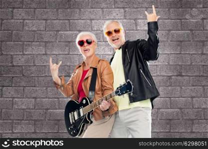 music, age and people concept - happy senior couple in sunglasses with electric guitar showing rock hand sign over brick wall background. senior couple with guitar showing rock hand sign