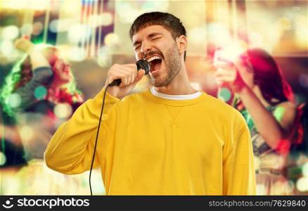 music and people concept - young man in yellow sweatshirt with microphone singing karaoke over night club background. man with microphone singing at night club