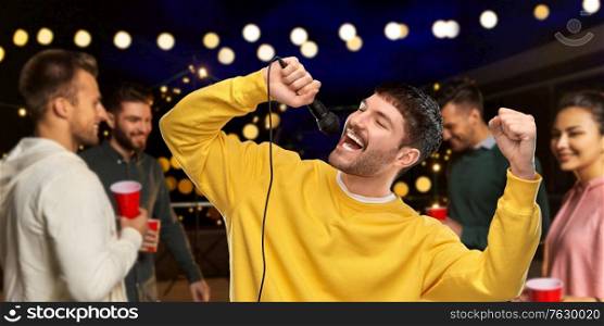 music and people concept - young man in yellow sweatshirt with microphone singing karaoke over night rooftop party background. man with microphone singing at night rooftop party