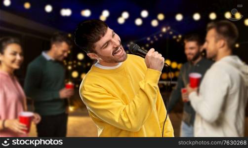 music and people concept - young man in yellow sweatshirt with microphone singing karaoke over night rooftop party background. man with microphone singing at night rooftop party