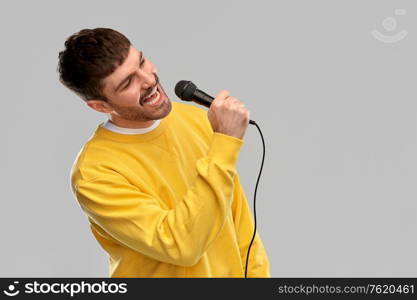 music and people concept - young man in yellow sweatshirt with microphone singing over grey background. man in yellow sweatshirt with microphone singing