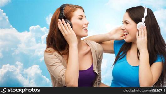 music and technology concept - two laughing teenage girls or young women with headphones listening to music over blue sky with clouds background. two happy girls with headphones listening to music