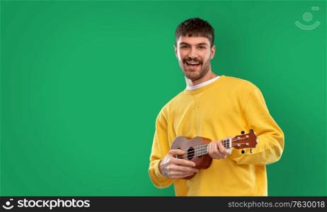 music, leisure and people concept - smiling young man in yellow sweatshirt playing ukulele guitar over emerald green background. happy man playing ukulele guitar over green