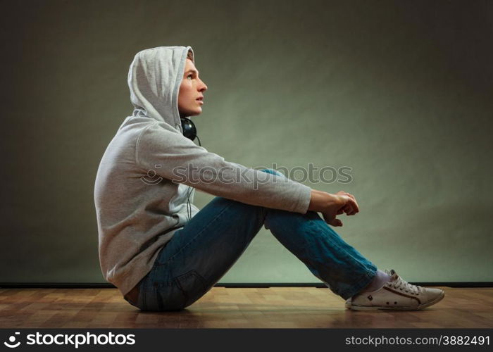 Music passion youth concept. Serious hooded man teen boy with headphones sitting daydreaming on floor grunge background