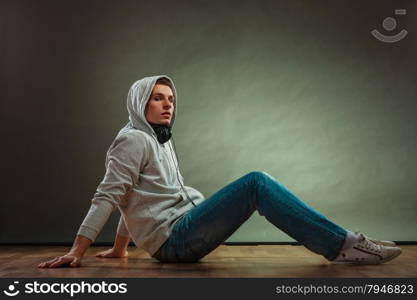 Music passion youth concept. Serious hooded man teen boy with headphones sitting daydreaming on floor grunge background