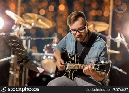 music, people and entertainment concept - male guitarist playing electric guitar at studio rehearsal over holidays lights background. musician playing guitar at studio or music concert