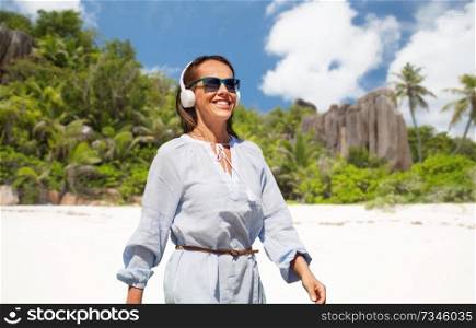 music, people and technology concept - happy smiling woman with headphones walking over tropical beach on seychelles island background. woman with headphones walking along summer beach