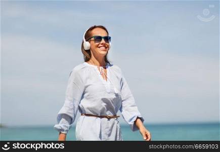 music, people and technology concept - happy smiling woman with headphones walking along summer beach. woman with headphones walking along summer beach
