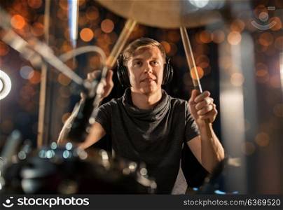 music, people, musical instruments and entertainment concept - male musician in headphones with drumsticks playing drum kit at concert or studio over holidays lights background. male musician playing drum kit at concert