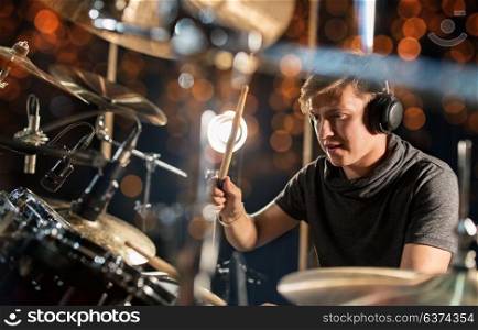 music, people, musical instruments and entertainment concept - male musician in headphones with drumsticks playing drum kit at concert or studio over holidays lights background. male musician playing drum kit at concert