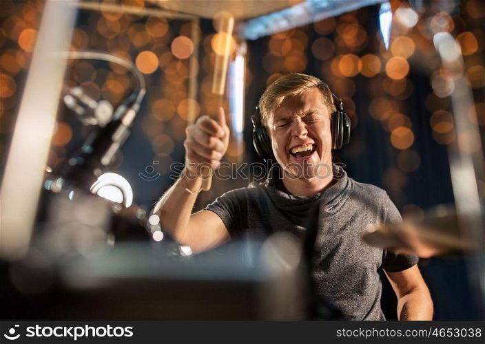 music, people, musical instruments and entertainment concept - male musician or drummer in headphones with drumsticks playing drums and cymbals at concert or studio over holidays lights background. musician playing drum kit at concert over lights