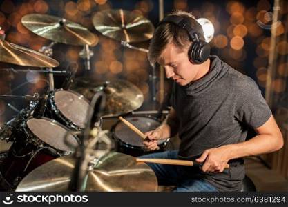 music, people, musical instruments and entertainment concept - male musician or drummer in headphones with drumsticks playing drums and cymbals at concert or studio over holidays lights background. musician playing drum kit at concert over lights