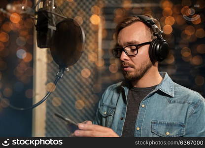 music, show business, people and technology concept - male singer with headphones, microphone and smartphone at sound recording studio over holidays lights background. singer with headphones at music recording studio