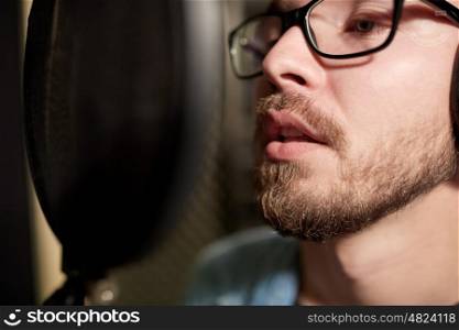 music, show business, people and voice concept - close up of male singer with headphones and microphone singing song at sound recording studio