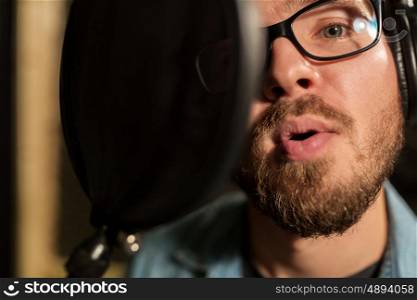music, show business, people and voice concept - close up of male singer with headphones and microphone singing song at sound recording studio