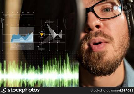 music, show business, people and voice concept - close up of male singer with headphones and microphone singing song at sound recording studio. man singing at sound recording studio