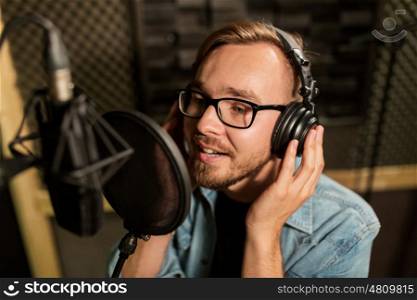 music, show business, people and voice concept - male singer with headphones and microphone singing song at sound recording studio