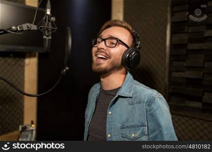 music, show business, people and voice concept - male singer with headphones and microphone singing song at sound recording studio