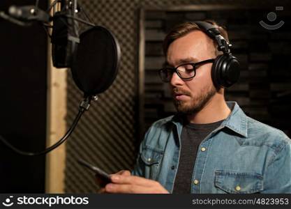 music, show business, people and voice concept - male singer with headphones, microphone and smartphone at sound recording studio
