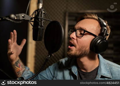 music, show business, people and voice concept - male singer with headphones and microphone singing song at sound recording studio