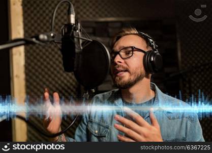music, show business, people and voice concept - male singer with headphones and microphone singing song at sound recording studio. man with headphones singing at recording studio